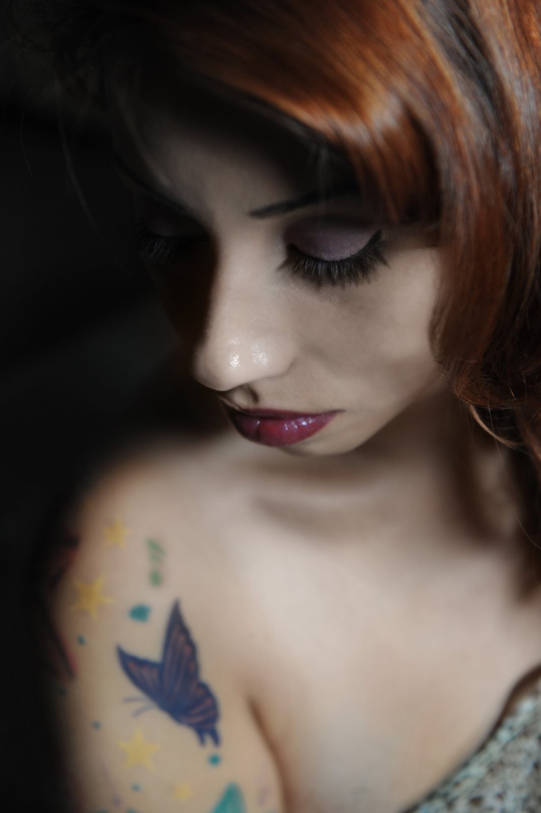 Close portrait of a woman with auburn hair and purple eye makeup looking down, a butterfly tattoo on her shoulder