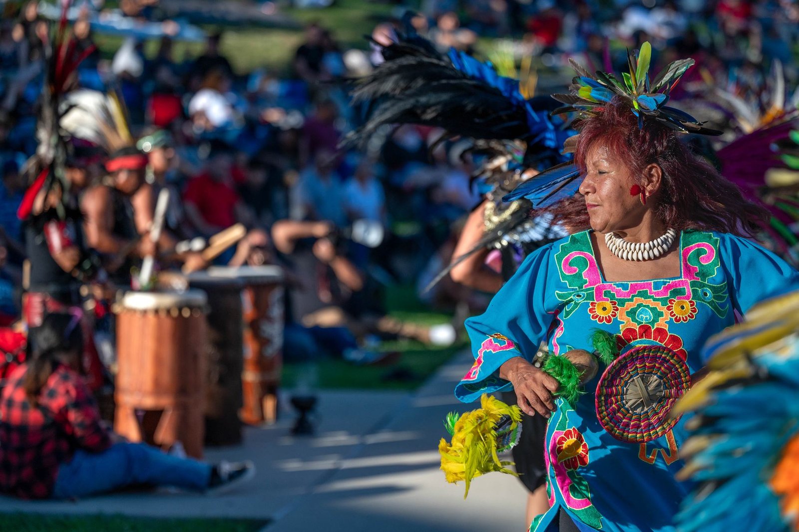 A woman in a bright blue embroidered blouse and a feathered headdress dances in a park while drummers and a crowd sit behind her