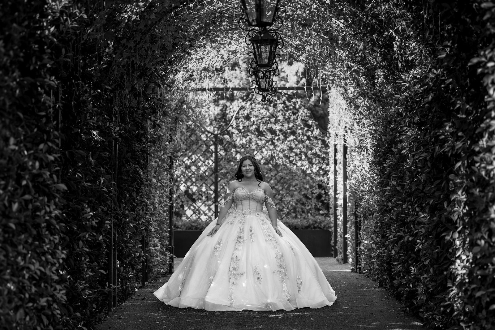 Black and white photograph of a young woman in a full ball gown standing at the end of a tunnel of ivy with a lantern hanging overhead