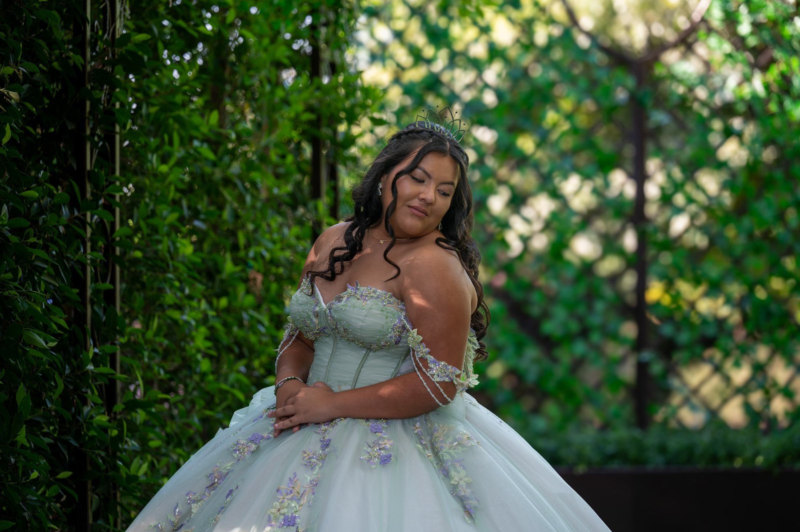 A young woman in an off the shoulder sage green gown and a tiara looks down and to the side in a garden, framed by green hedges