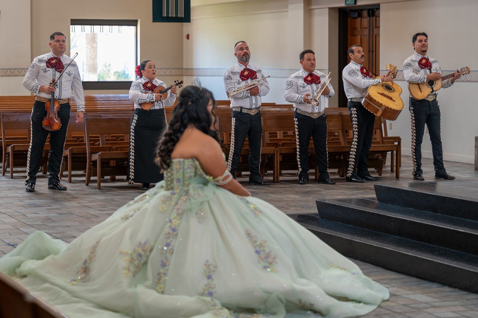 A young woman in a sage green ball gown kneels in a church aisle facing a mariachi band in white jackets