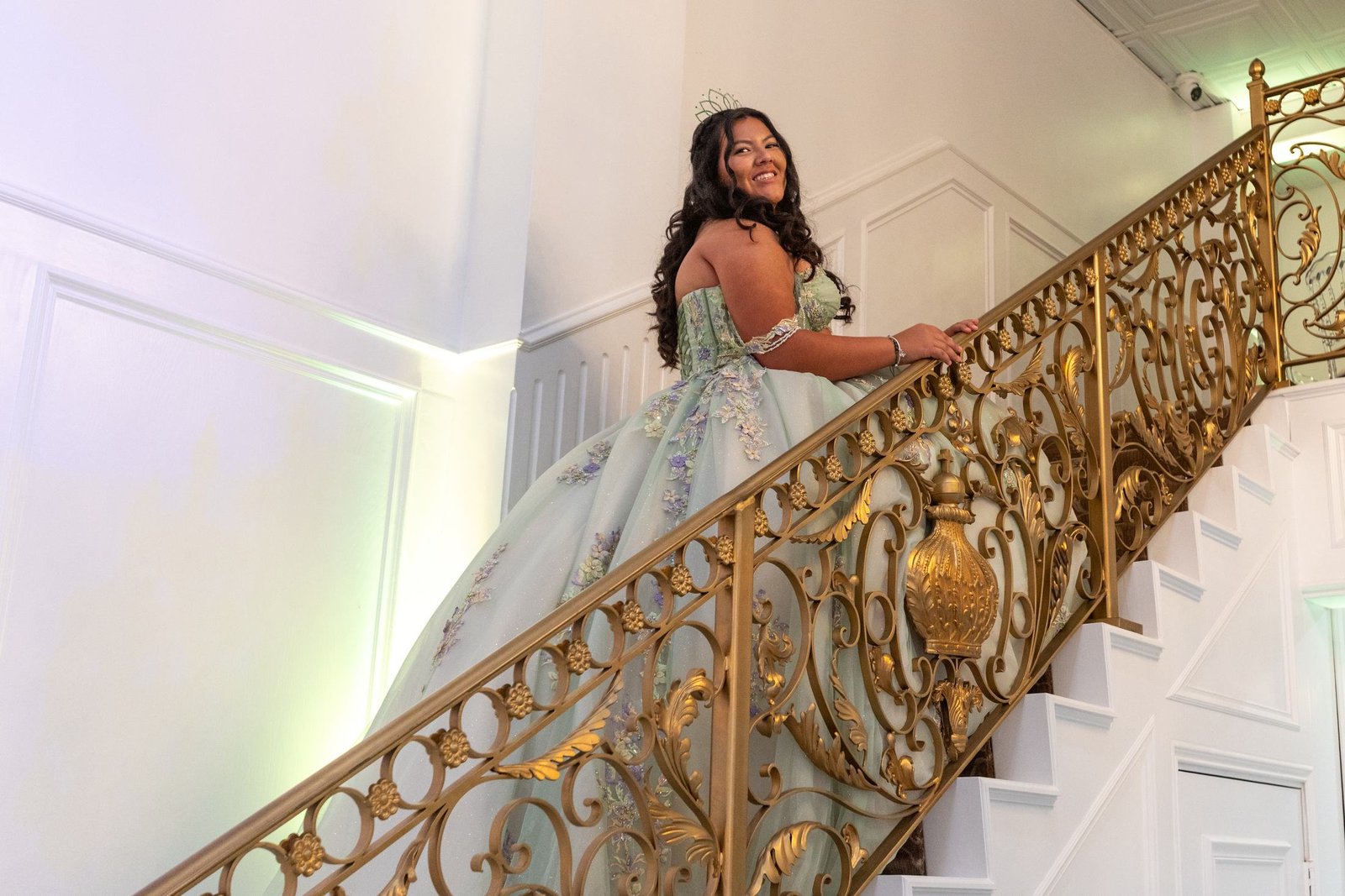 A young woman in a sage green gown and a tiara climbs a white staircase with an ornate gold railing and looks back over her shoulder