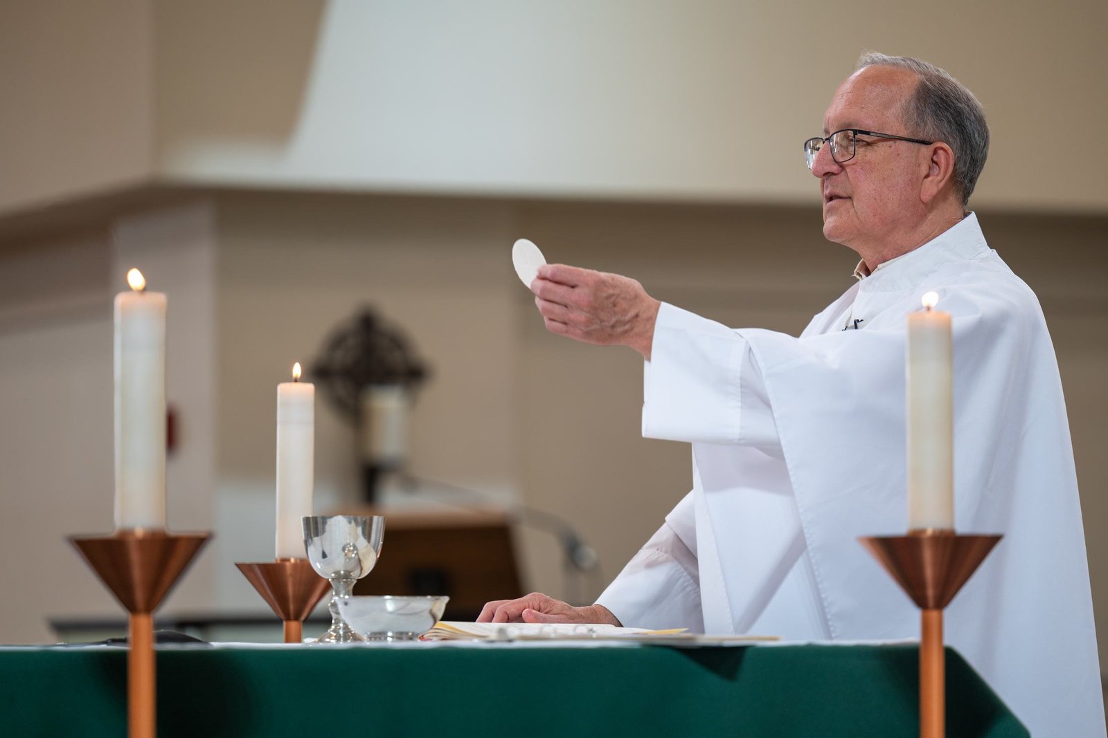 A priest in white vestments holds up a communion wafer at an altar between two tall candles
