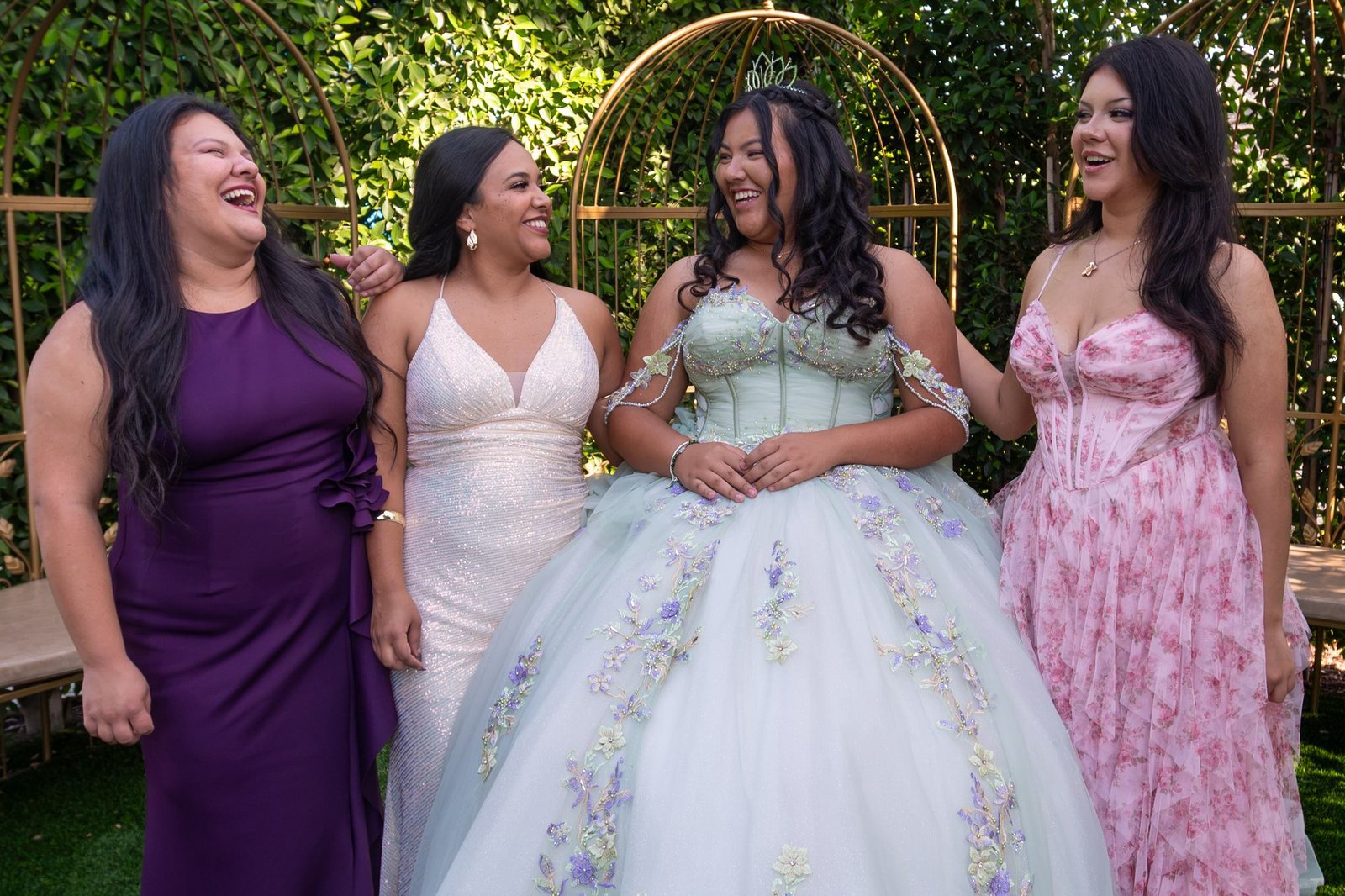 A young woman in a sage green gown and a tiara laughs with three women in a garden, wearing purple, white and pink