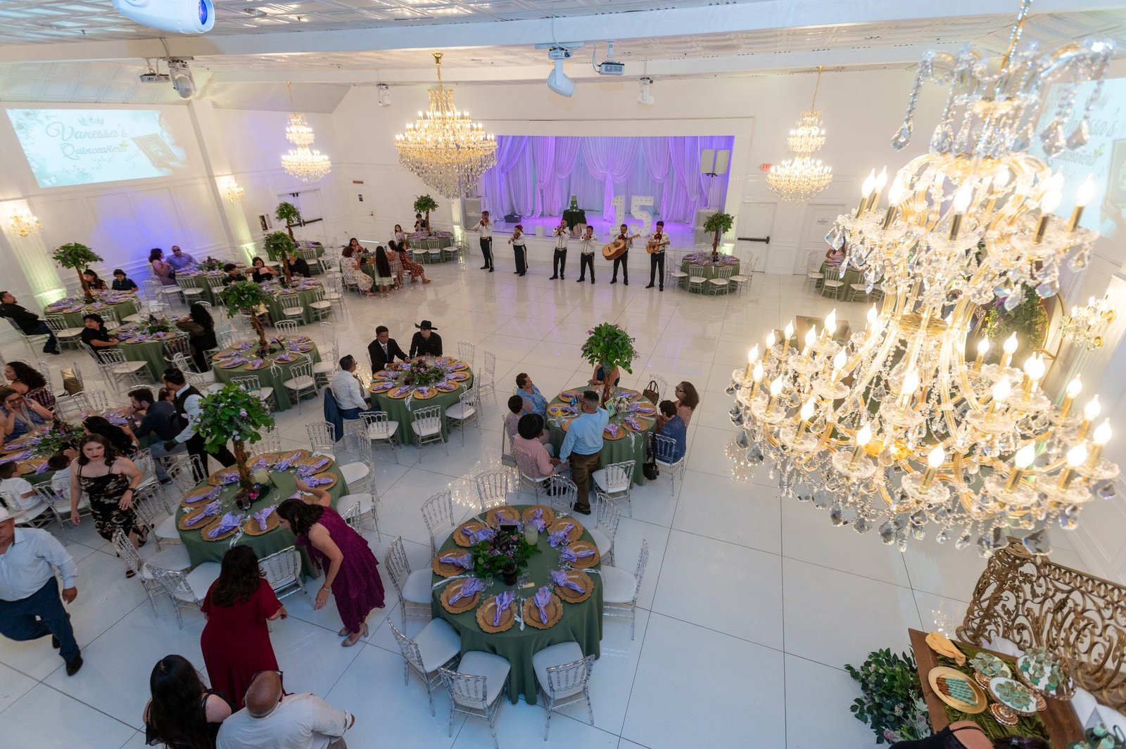 High angle view of a banquet hall with a white floor, round tables set in sage green, guests, a mariachi group and large crystal chandeliers