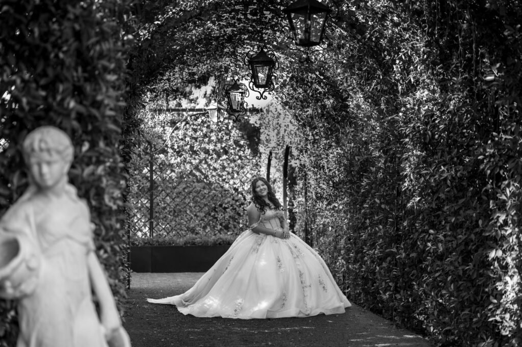 Black and white photograph of a young woman in a ball gown seated on a garden path beneath an ivy covered arch