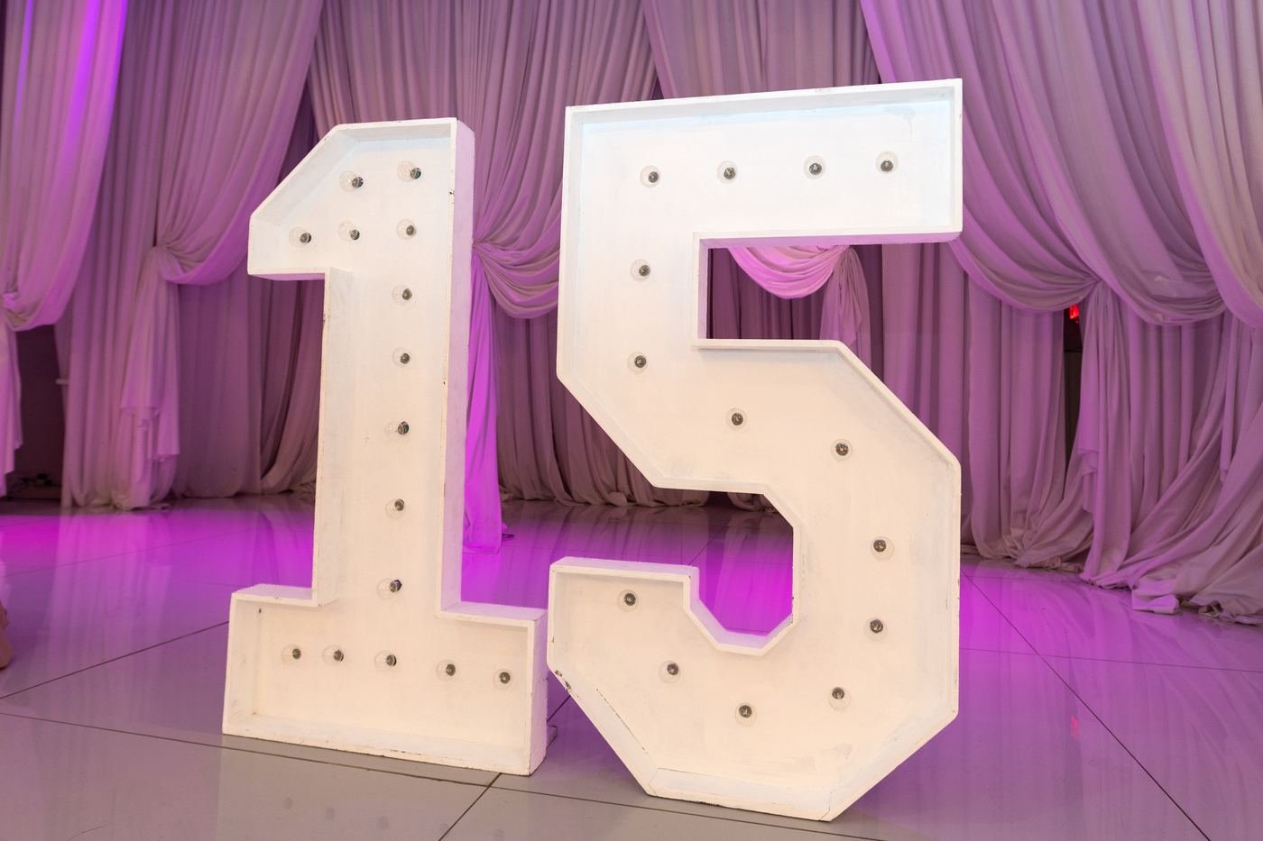A lit marquee sign spelling 15 in front of draped lavender curtains and a glowing dance floor