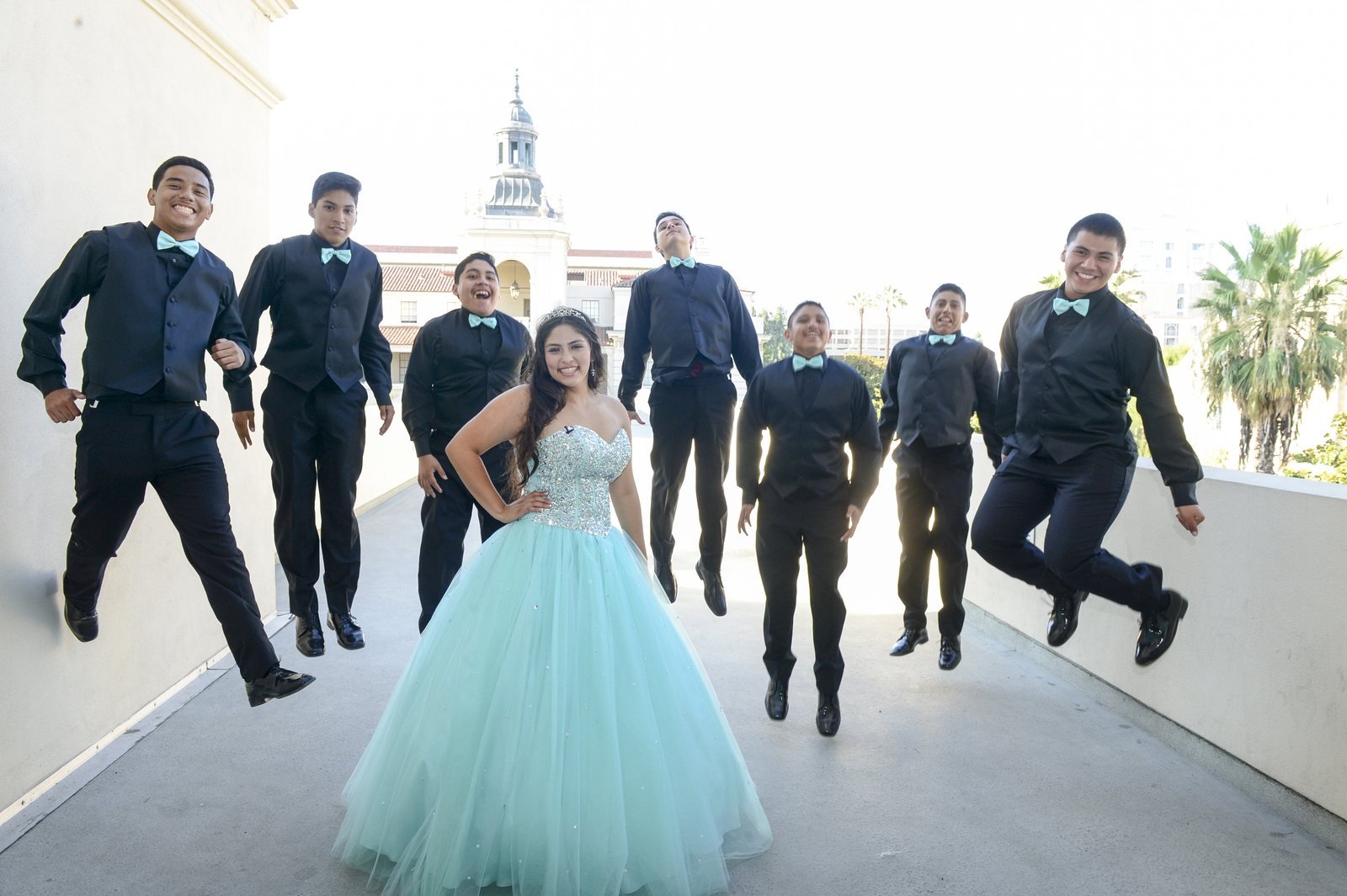 A young woman in an aqua ball gown poses on a sunlit balcony while seven young men in black vests and aqua bow ties jump behind her