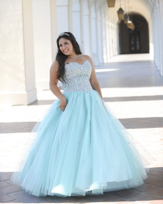 A smiling young woman in a crystal bodice and aqua ball gown stands in a sunlit arched corridor, wearing a tiara
