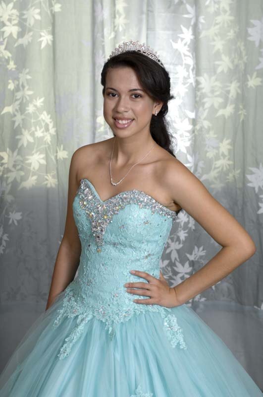 A smiling young woman in a tiara and aqua lace gown in front of a sheer silver curtain