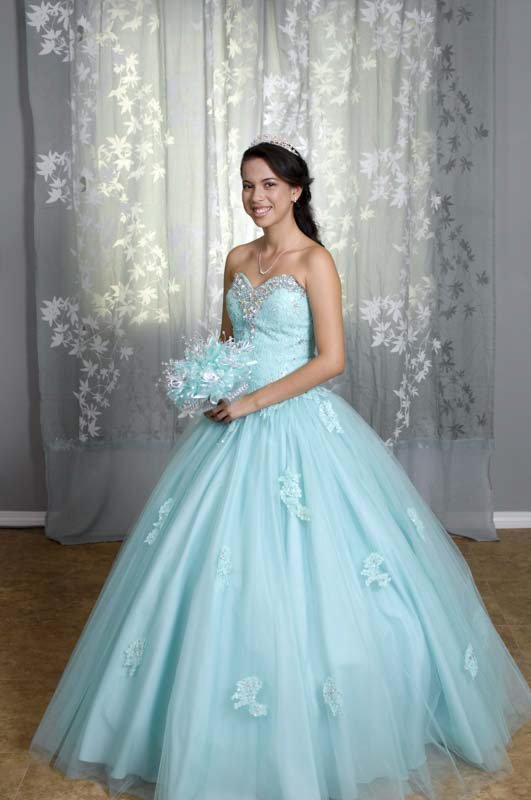 A young woman in a tiara and a full aqua gown with lace appliques holds a bouquet of pale blue flowers in front of a sheer curtain